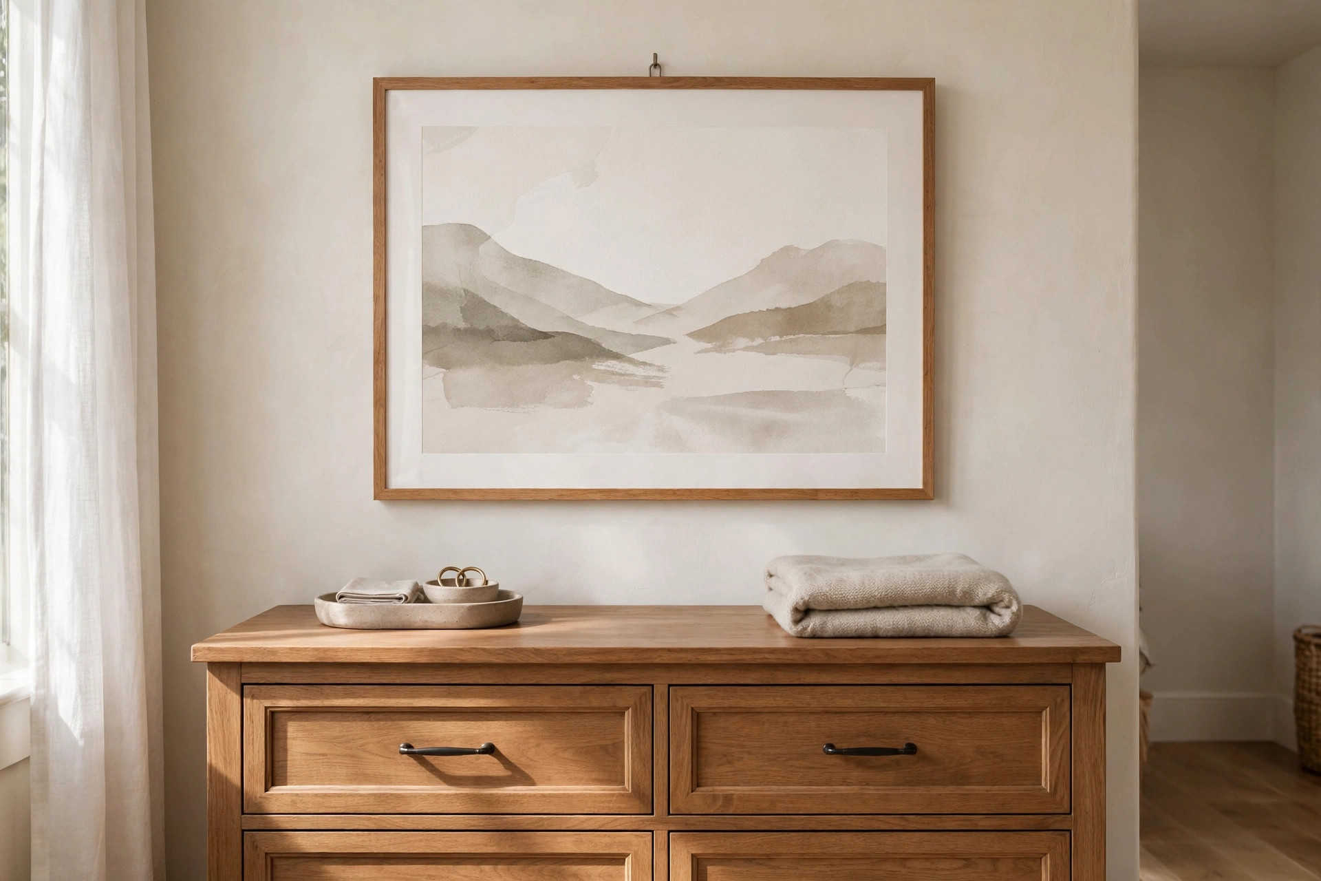 Large soft watercolor landscape in a wood frame centered above a wooden dresser with a tray and folded blanket