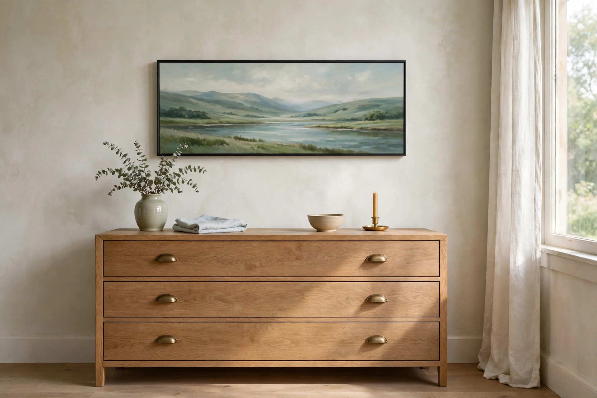Low wide landscape painting of a river and hills in a thin black frame above a wooden dresser