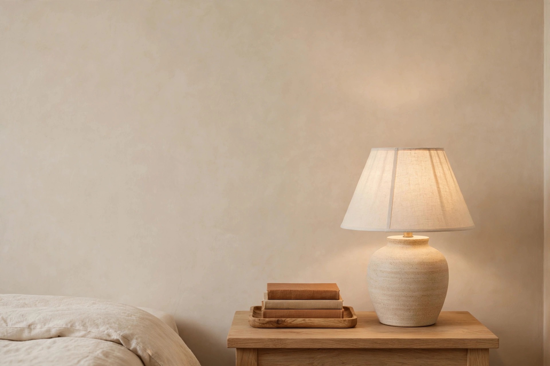 Wood nightstand with a matte ceramic lamp and a wood tray holding warm toned books beside a cream bed