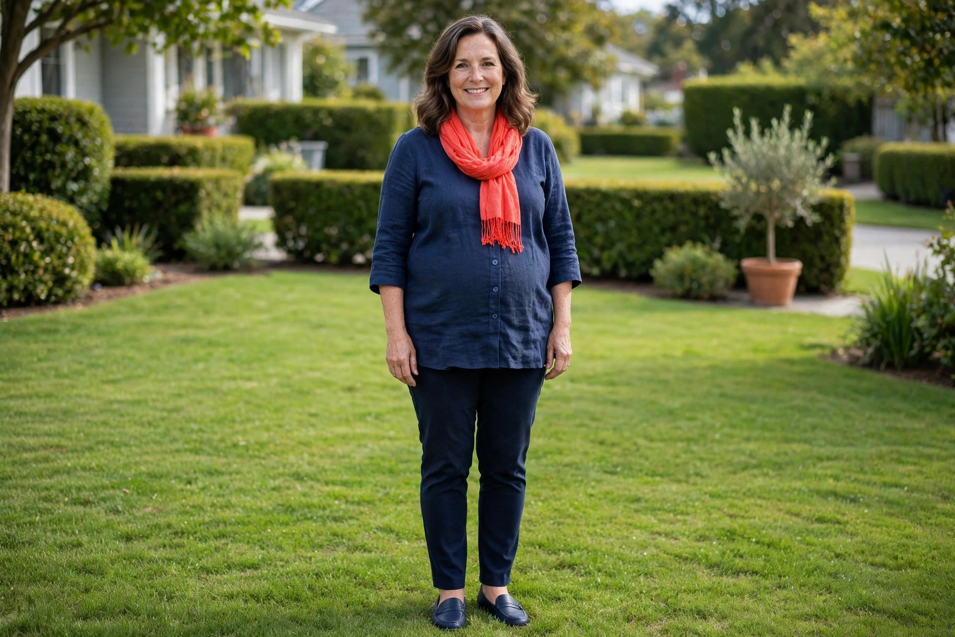 Woman wearing a navy tunic, navy slim pants and navy loafers with a coral scarf on a green lawn