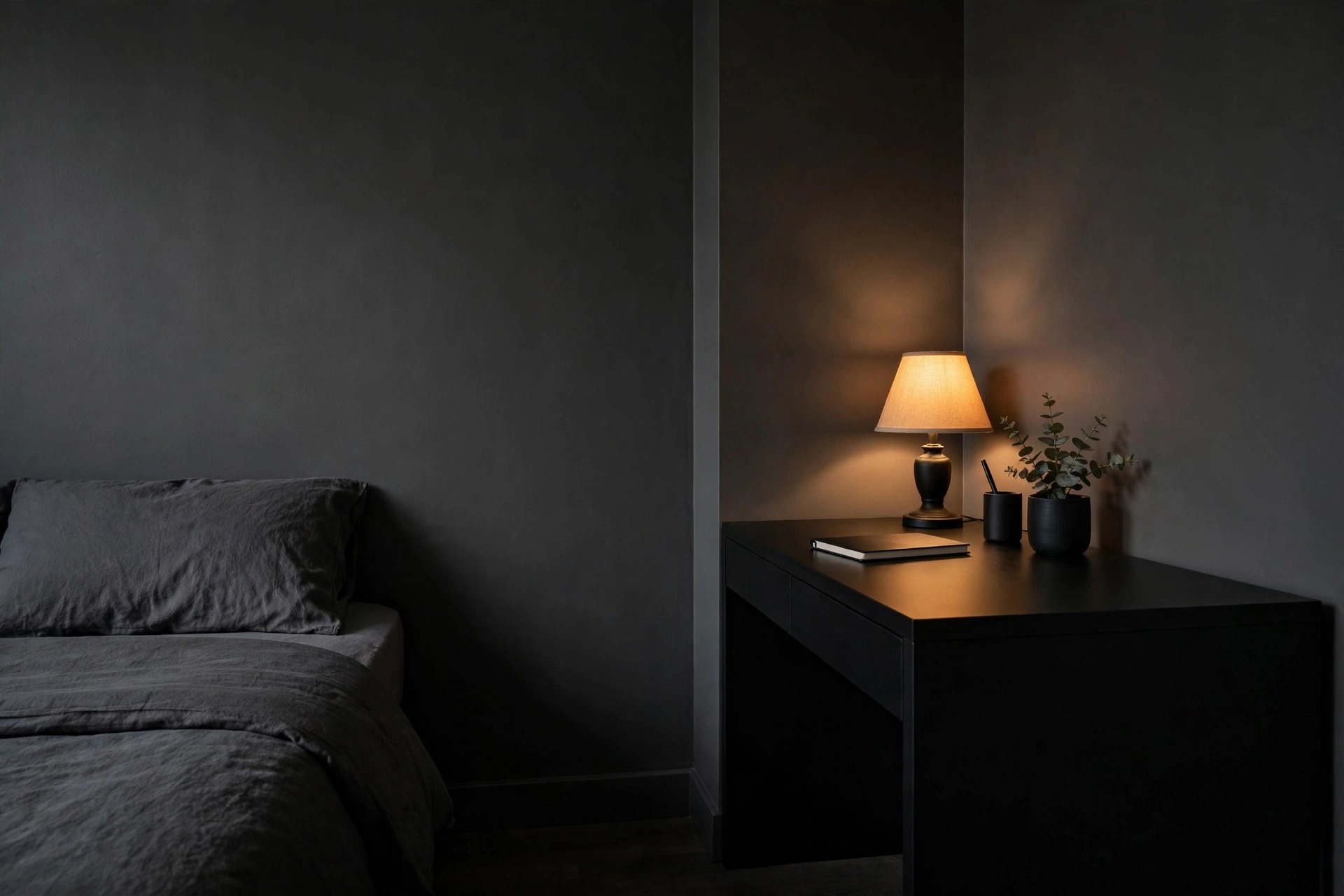Dark teen bedroom corner with a matte charcoal wall and a matte black desk in flat finishes, lit by one warm table lamp, with the edge of a bed with dark bedding visible