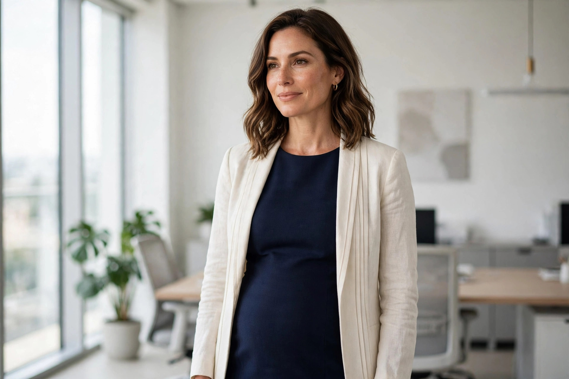 Woman wearing a navy column dress with an open cream blazer in a bright modern office