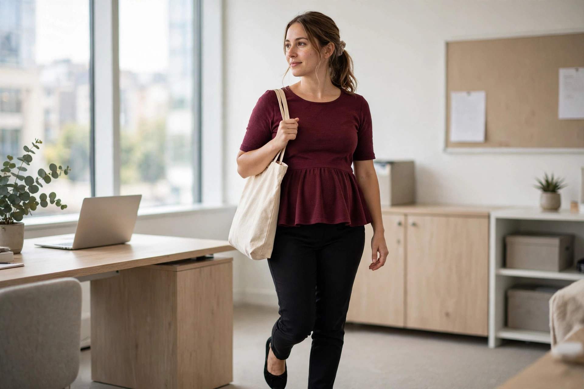 Woman in a burgundy peplum top and black slim ankle trousers carrying a tote in an office