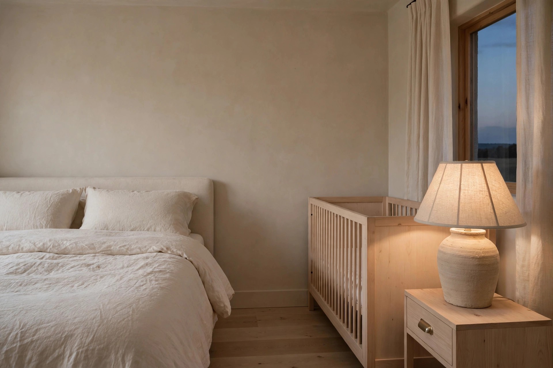 Dusk beige bedroom with an unglazed ceramic lamp with a fabric shade on a light wood nightstand beside a raw wood crib and a cream bed