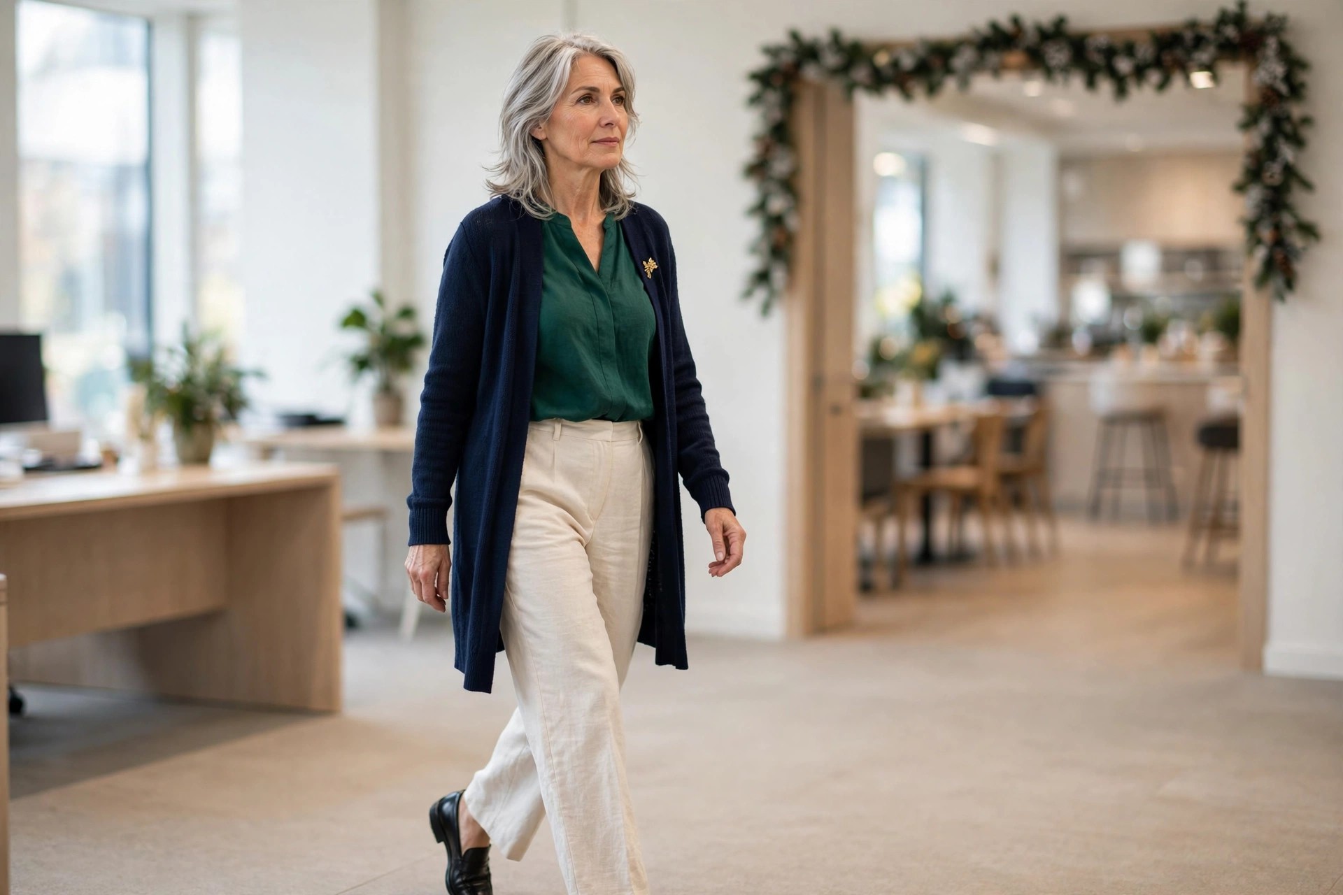 Woman over 50 with silver hair wearing cream trousers, an emerald blouse and a navy long cardigan with a gold brooch walking through an office